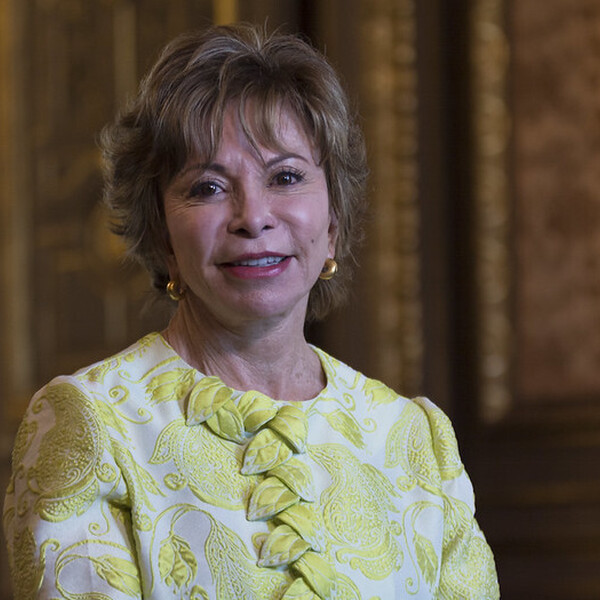 A headshot of Isabel Allende. She has short curly hair and is wearing a yellow blazer.