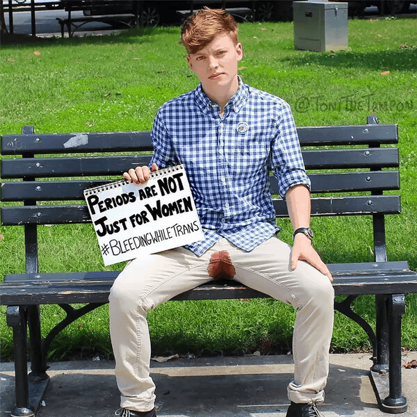 A trans and non-binary person sitting on a bench bleeding through their pants as they hold a sign that says, "Periods are not just for women. #BleedingWhileTrans."