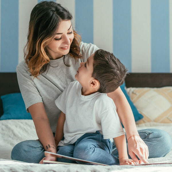 A mother and son sit on a bed together with a book in front of them.