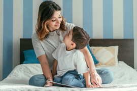 A mother and son sit on a bed together with a book in front of them.