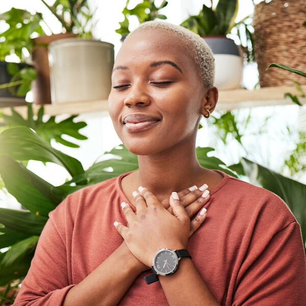 A black woman stands in front of plants with her eyes closed and her hands over her chest