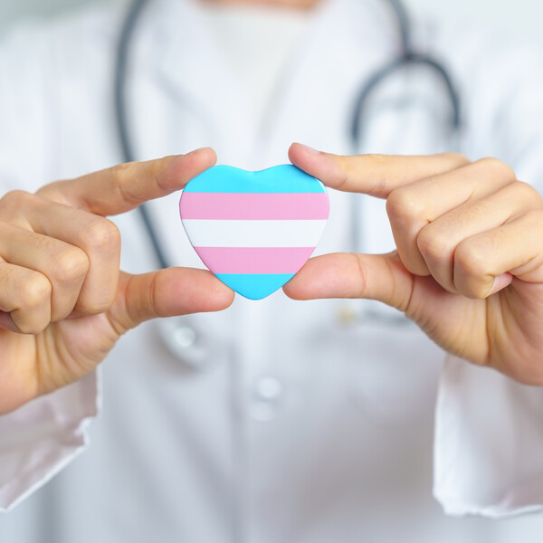 A photo of a white doctor with a stethoscope around their neck holding a heart with a trans flag
