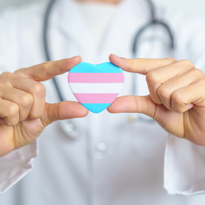 A photo of a white doctor with a stethoscope around their neck holding a heart with a trans flag