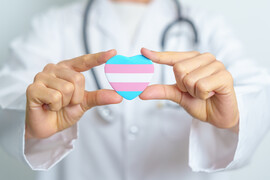 A photo of a white doctor with a stethoscope around their neck holding a heart with a trans flag