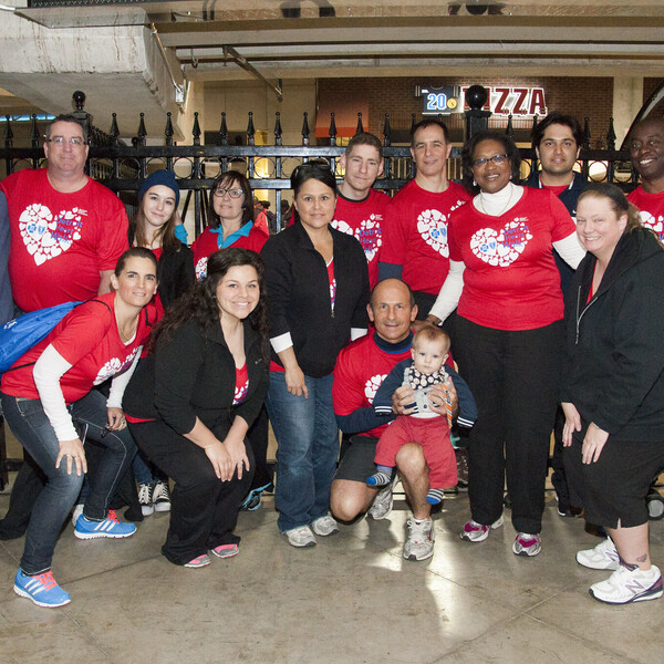 A large group of people, many wearing red shirts with white hearts on them, stand in front of a black fence.