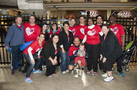 A large group of people, many wearing red shirts with white hearts on them, stand in front of a black fence.