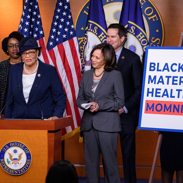 A Black woman, Alma Adams, stands at a podium at the U.S. House of Representatives. She is flanked by a group of people, including Kamala Harris. A poster near her reads "Black Maternal Health Momnibus."