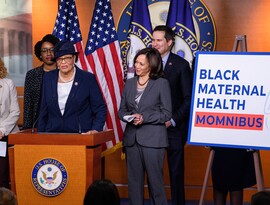 A Black woman, Alma Adams, stands at a podium at the U.S. House of Representatives. She is flanked by a group of people, including Kamala Harris. A poster near her reads "Black Maternal Health Momnibus."