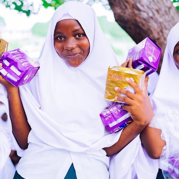 Two smiling girls wearing white headscarves hold menstrual products in their hands.