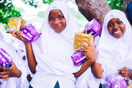 Two smiling girls wearing white headscarves hold menstrual products in their hands.