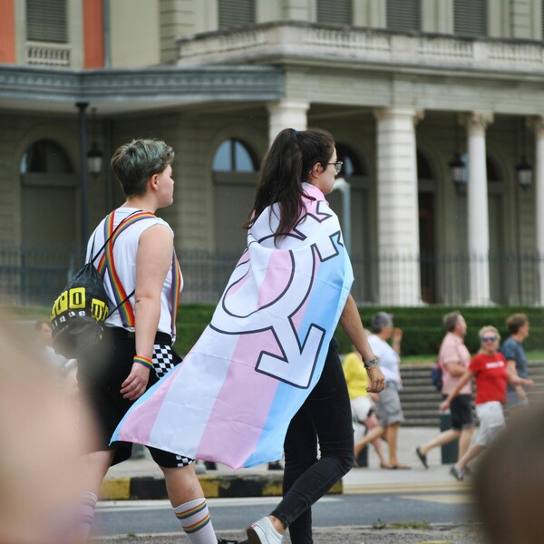 Two people, one with short hair and one wrapped in a trans pride flag, walking on road at a pride parade in Geneva.