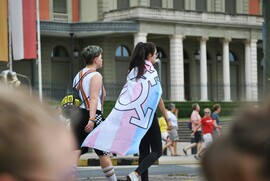 Two people, one with short hair and one wrapped in a trans pride flag, walking on road at a pride parade in Geneva.