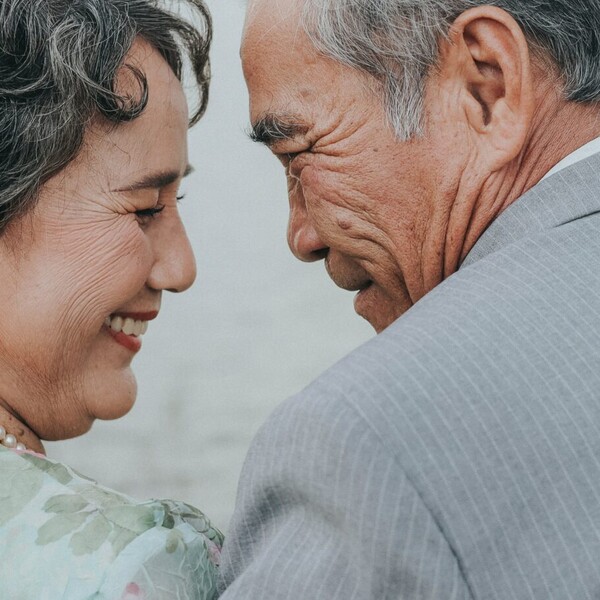An older Asian couple smile as they face each other. They have gray hair. He is wearing a gray suit, and she is wearing a green dress with a pearl necklace. The image is taken from behind them.