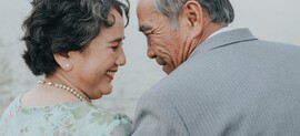 An older Asian couple smile as they face each other. They have gray hair. He is wearing a gray suit, and she is wearing a green dress with a pearl necklace. The image is taken from behind them.