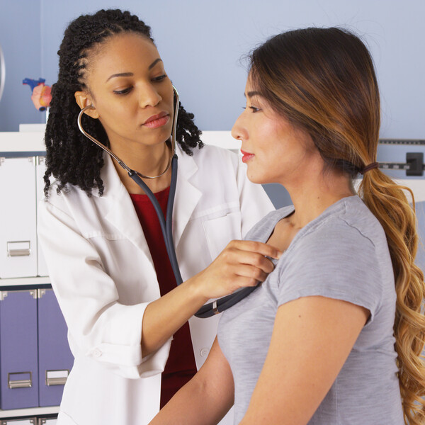 A Black doctor in a white coat and her hair styled in twists listens to a Latina woman's heart beat in a doctor's office