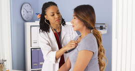 A Black doctor in a white coat and her hair styled in twists listens to a Latina woman's heart beat in a doctor's office