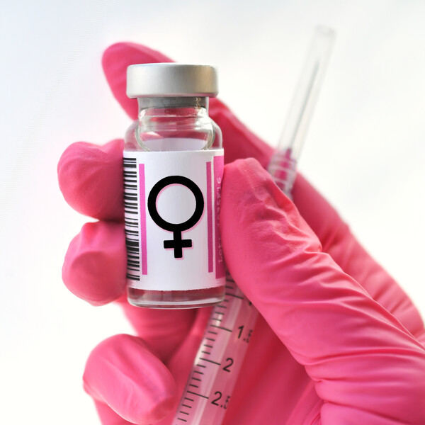 A white background. A hand with a pink glove holding a vial and needle with the female symbol.