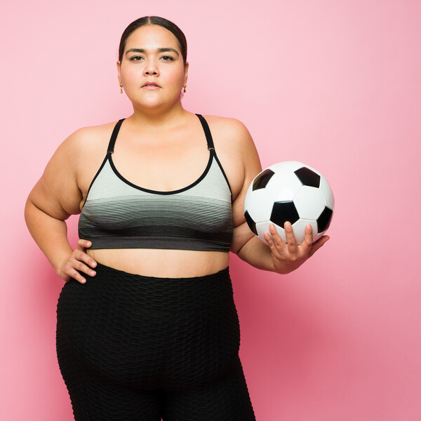 A pink background behind a fat Latina woman wearing a sports bra and leggings. She is holding a soccer ball.