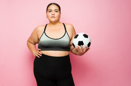 A pink background behind a fat Latina woman wearing a sports bra and leggings. She is holding a soccer ball.