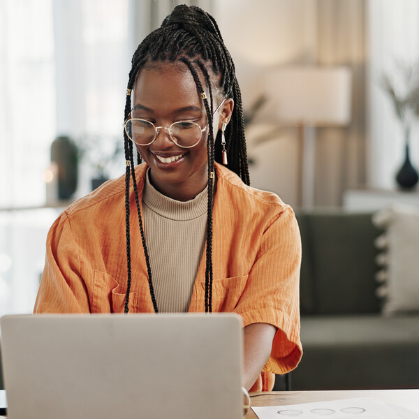 A Black woman sits in what appears to be her living room. She is working on a gold mac. Her hair is styled in braids pulled up, she is wearing glasses, and she is wearing an orange top.