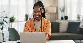 A Black woman sits in what appears to be her living room. She is working on a gold mac. Her hair is styled in braids pulled up, she is wearing glasses, and she is wearing an orange top.