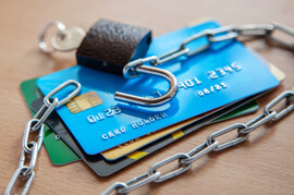 A stack of credit cards. From the bottom, they are green, black, gold, and blue. On top of them is a lock with a chain attached.