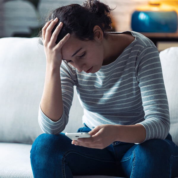A white woman in a striped gray shirt looks down at her phone as she sits on the couch. Her hand is on her head as though she is concerned.