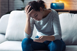 A white woman in a striped gray shirt looks down at her phone as she sits on the couch. Her hand is on her head as though she is concerned.