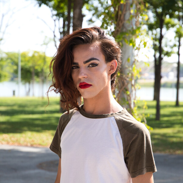 A trans woman stands in a park. She is wearing a green and white t-shirt and red lipstick. She has red hair that is pulled to one side.