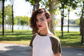 A trans woman stands in a park. She is wearing a green and white t-shirt and red lipstick. She has red hair that is pulled to one side.