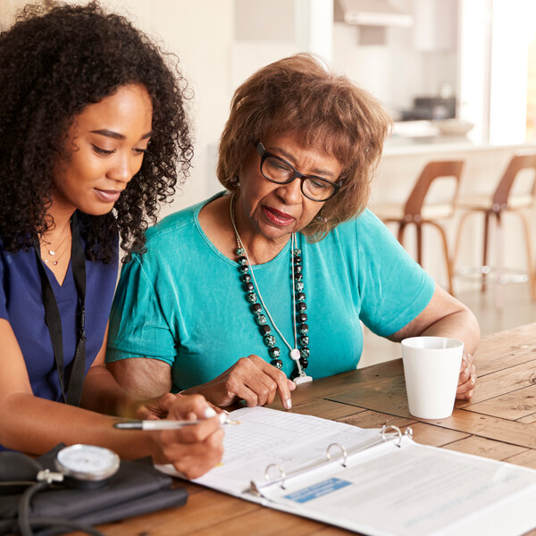 An older Black woman with brown hair wearing a blue top and glasses sits next to a Black woman home aide nurse with curly hair who is filling out paperwork for the older woman.