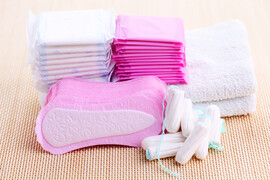 A stack of pink pads next to a stack of white pads. Next to them are white towels. In front of those stacks are pink panty liners and white tampons.