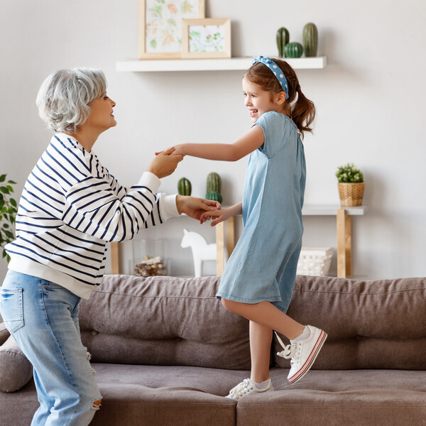 An older woman in a gray sweater and jeans with gray hair dances with her granddaughter who is standing on the couch in the living room