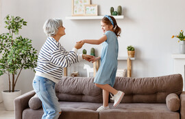 An older woman in a gray sweater and jeans with gray hair dances with her granddaughter who is standing on the couch in the living room