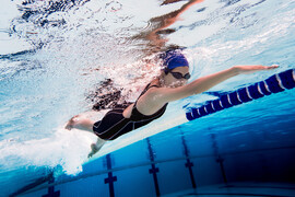 A woman in a black swimsuit, black goggles, and navy swim cap swims in a swimming pool