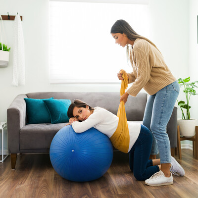A Latina woman doula in a sweater and jeans lifts a pregnant woman's belly with a yellow rebozo, a Mexican garment. The pregnant woman is lying on a blue exercise ball and is wearing a white shirt and jeans.