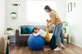 A Latina woman doula in a sweater and jeans lifts a pregnant woman's belly with a yellow rebozo, a Mexican garment. The pregnant woman is lying on a blue exercise ball and is wearing a white shirt and jeans.