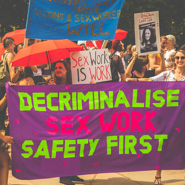 A protest. Centered is a large purple sign that says, "Decriminalize Sex Worker Safety First" in hot pink and highlighter green. The sign is held by 3 women. Behind are people holding other signs that read: "Sex Work is Work" and "Stop Police Raids. Consenting Sex is Not a Crime!" Some people standing around are holding red umbrellas.