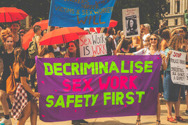 A protest. Centered is a large purple sign that says, "Decriminalize Sex Worker Safety First" in hot pink and highlighter green. The sign is held by 3 women. Behind are people holding other signs that read: "Sex Work is Work" and "Stop Police Raids. Consenting Sex is Not a Crime!" Some people standing around are holding red umbrellas.