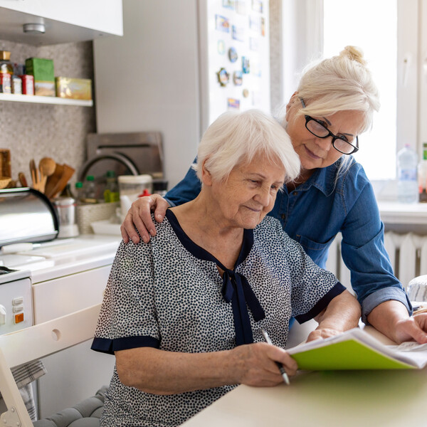 An older woman in a black and white top sits at a table in a kitchen holding folders. A younger woman in glasses and a jean shirt is holding the papers with her and standing next to her. They seem to be working on the papers together.