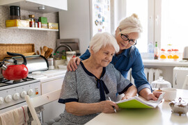 An older woman in a black and white top sits at a table in a kitchen holding folders. A younger woman in glasses and a jean shirt is holding the papers with her and standing next to her. They seem to be working on the papers together.