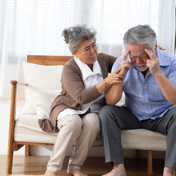 A senior Asian couple sitting on a couch together. The man has his hands on his head and the woman is tending to him.