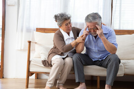 A senior Asian couple sitting on a couch together. The man has his hands on his head and the woman is tending to him.