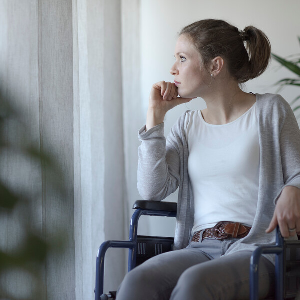 A woman sits in a wheelchair facing the camera and looks out the window. Her hair is pulled back into a ponytail, and she is wearing a white shirt, gray sweater, and gray jeans.