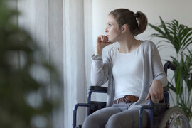 A woman sits in a wheelchair facing the camera and looks out the window. Her hair is pulled back into a ponytail, and she is wearing a white shirt, gray sweater, and gray jeans.