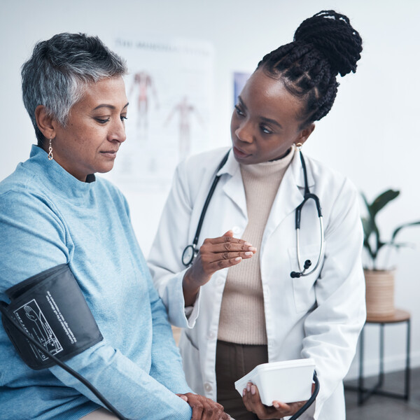 An older woman with short hair who appears to be South Asian and is wearing a blue shirt gets her blood pressure checked by a Black woman in a white coat. She has a stethoscope wrapped around her neck, and her hair is styled in braids.