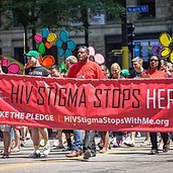 A group of people in a march. Many of them are wearing red t-shirts. Three people in the front are holding a red banner that says, "HIV Stigma Stops Here. Take the Pledge. HIVStigmaStopsWithMe.org."