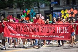 A group of people in a march. Many of them are wearing red t-shirts. Three people in the front are holding a red banner that says, "HIV Stigma Stops Here. Take the Pledge. HIVStigmaStopsWithMe.org."