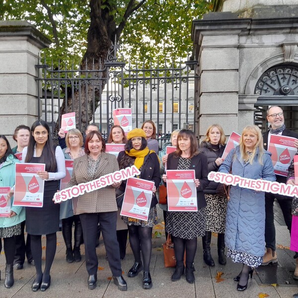 A group of women in coats and hats standing in front of a gated resident holding signs saying "Stop the Stigma."