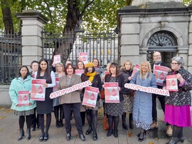 A group of women in coats and hats standing in front of a gated resident holding signs saying "Stop the Stigma."